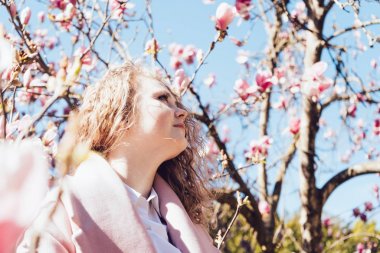 pembe bir ceket güzel kıvırcık kadın parkta yürüyor, güzel kokulu Manolya açık havada bahar güneş ışınlarının sahiptir