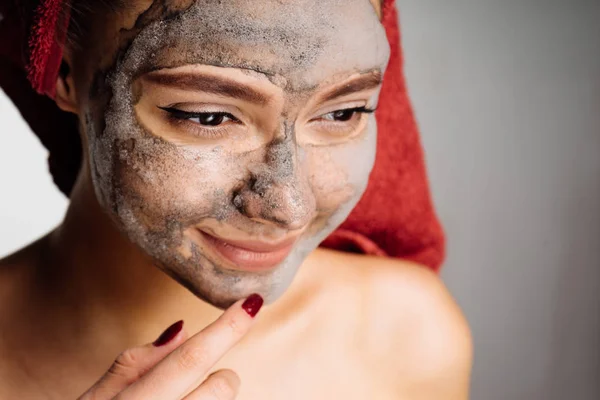 Belle jeune fille avec une serviette rouge sur la tête applique un masque d'argile utile sur le visage — Photo