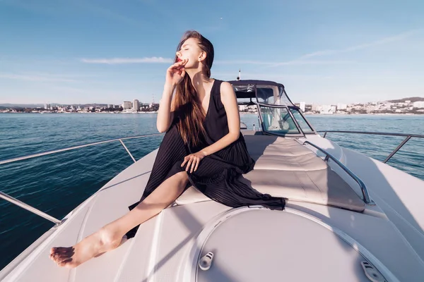 Una joven de lujo en un vestido negro se sienta en su gran yate blanco y disfruta de unas vacaciones en el mar —  Fotos de Stock