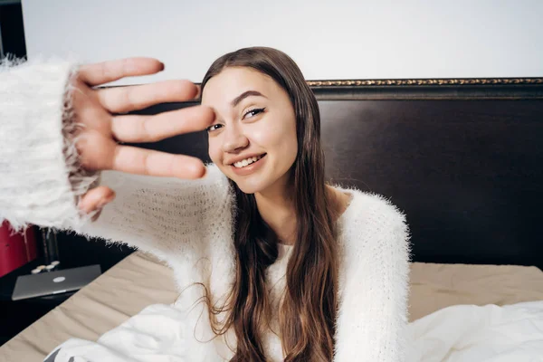 Lustiges junges Mädchen sitzt frühmorgens im Bett, lacht, schließt die Kamera mit der Hand — Stockfoto