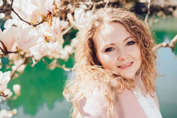 Cute curly-haired girl posing in the park in the rays of the spring sun, enjoying magnolia and a pond — Stock Photo, Image