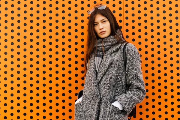 Fille de la ville aux cheveux foncés dans un manteau gris tendance posant contre un mur orange inhabituel — Photo