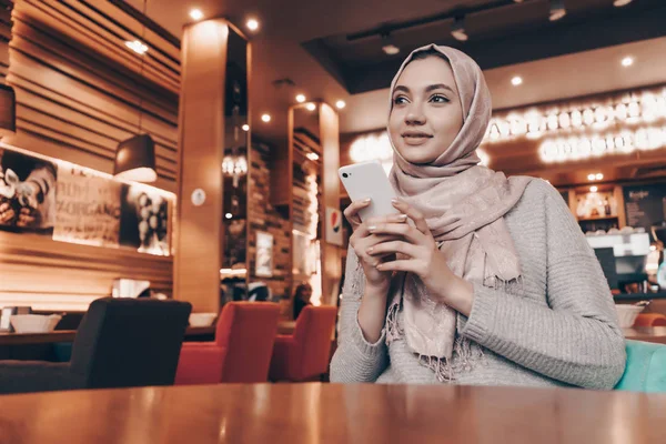 Schöne muslimische Mädchen mit Kopftuch sitzt auf Café, hält Smartphone in den Händen und lächelt — Stockfoto