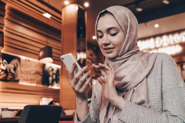 Милая арабская девушка с платком сидит в кафе и смотрит в ее смартфон, уютное место — стоковое фото