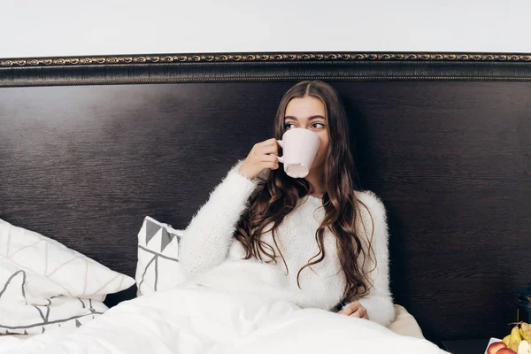 Långhåriga ung flicka sitter i sängen tidigt på morgonen, äter frukost och dricker doftande kaffe — Stockfoto