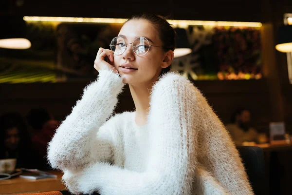 Молодая девушка в очках и белой куртке сидит в кафе после занятий — стоковое фото