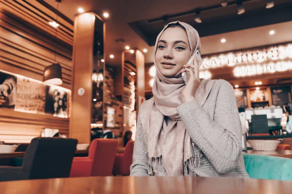 Busy young Muslim girl in hijab sitting in cafe and talking on the phone — Stock Photo, Image