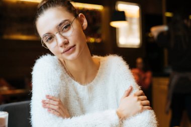 düşünceli genç kız serbest meslek ile gözlük ve beyaz bir kazak bir kafede oturuyor, kameraya benziyor