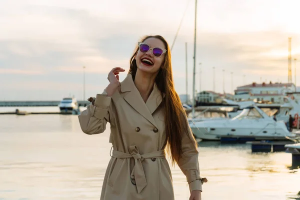 Szczęśliwa młoda kobieta w okularach słonecznych i modny beżowy płaszcz, ciesząc się na spacer nad rzeką — Zdjęcie stockowe