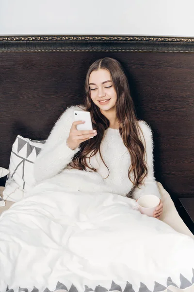 Красива довгошерста дівчина сидить у ліжку, відпочиває після важкого робочого дня, дивлячись в її смартфон і посміхаючись — стокове фото