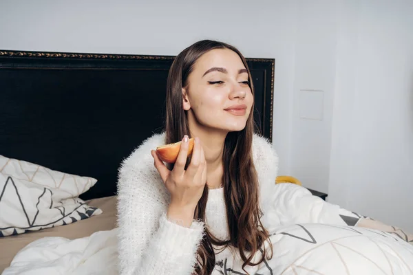 Divertida joven con el pelo largo sentado en la cama temprano en la mañana y tomar frutas del desayuno — Foto de Stock