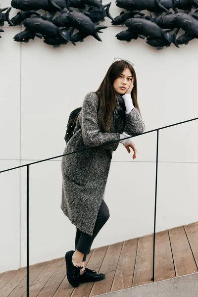 Dark-haired urban girl in a gray fashion coat is standing on the steps of an unusual building, street style — Stock Photo, Image