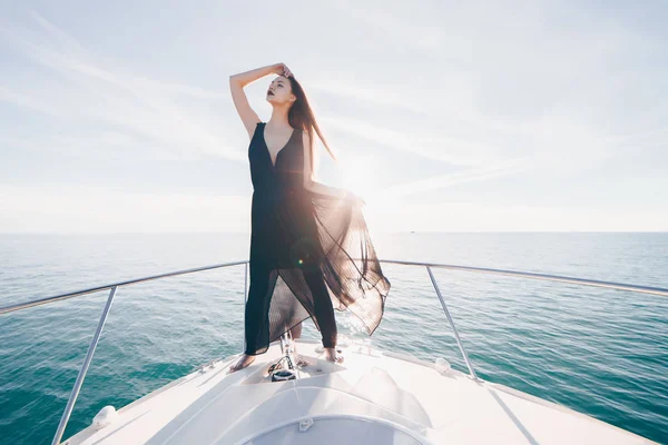 Una atractiva chica de lujo en un vestido negro está de pie en su yate blanco y disfrutar del sol y el mar, viajando —  Fotos de Stock