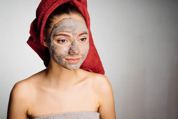 Hermosa joven con una toalla roja en la cabeza se aplica una máscara de arcilla útil en su cara, spa de día — Foto de Stock
