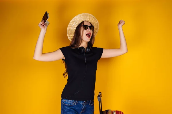 Feliz joven en gafas de sol y un sombrero va en un viaje, se regocija, con una gran maleta roja — Foto de Stock