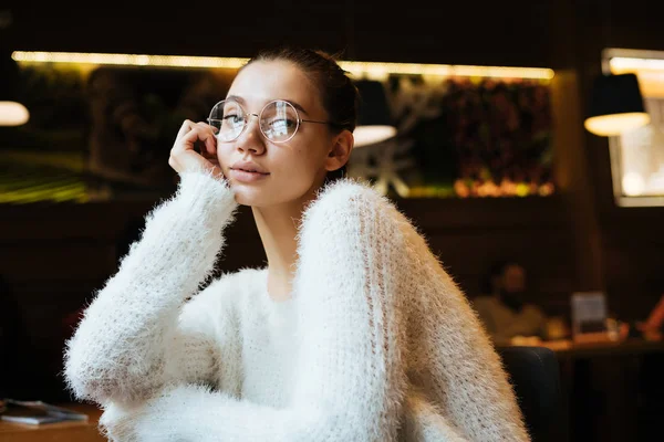 Красивая задумчивая юная студентка в очках и белый свитер сидит в кафе и смотрит в камеру — стоковое фото