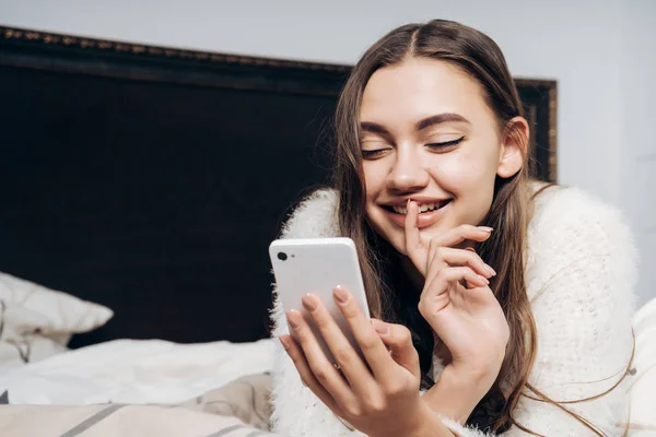 Красивая молодая девушка лежит в постели после рабочего дня, смотрит в свой смартфон и смеется — стоковое фото