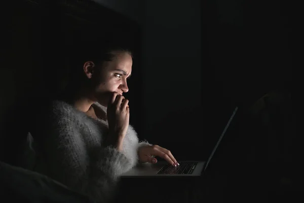 Сфокусированная задумчивая девушка, сидящая ночью в темноте и работающая на ноутбуке — стоковое фото