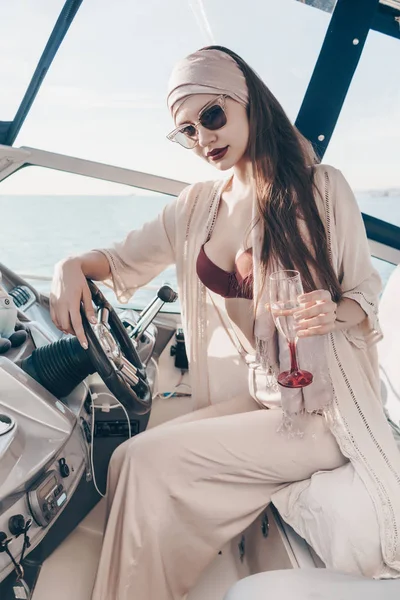 Una mujer joven rica y lujosa en gafas de sol y ropa elegante corre su yate blanco y bebe champán —  Fotos de Stock