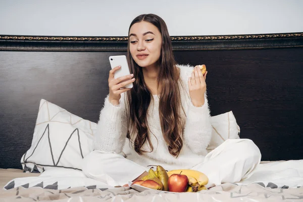 Niña feliz en pijama sentada en la cama temprano en la mañana, comiendo fruta, mirando el teléfono inteligente y riendo — Foto de Stock