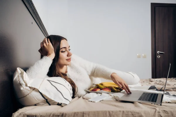 Nettes junges Mädchen liegt abends nach der Arbeit im Bett, schaut Fernsehserien auf Laptop und isst Obst — Stockfoto