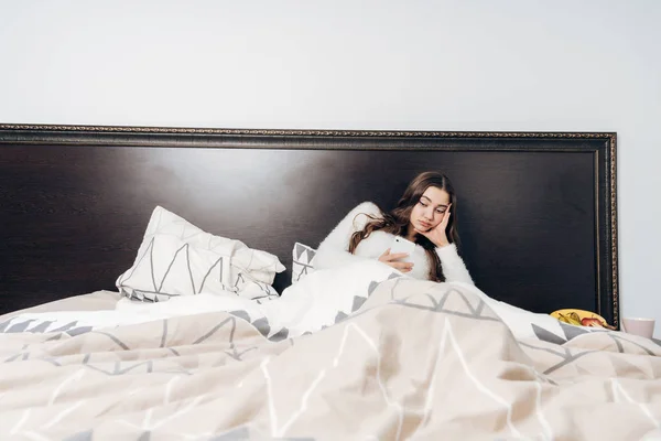 疲れた少女は仕事の後ベッドで横になっている彼女のスマート フォンに見えます、寝たいです。 — ストック写真