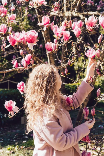 Mujer joven y rizada caminando en el parque en un día soleado, de pie junto a una magnolia rosa — Foto de Stock