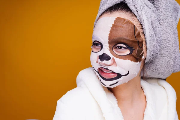 Divertente ragazza con un asciugamano sulla testa, ridendo, sulla maschera facciale con il viso di un cane — Foto Stock