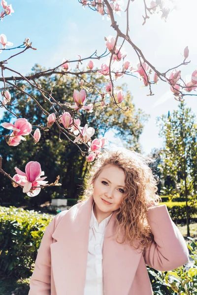 Приваблива кучерява молода жінка в рожевому пальто позує в парку в променях весняного сонця — стокове фото