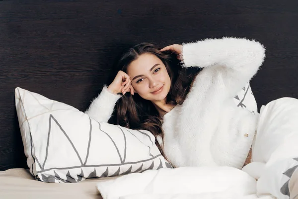Happy young girl in white jacket is lying in bed, resting after work and smiling — Stock Photo, Image
