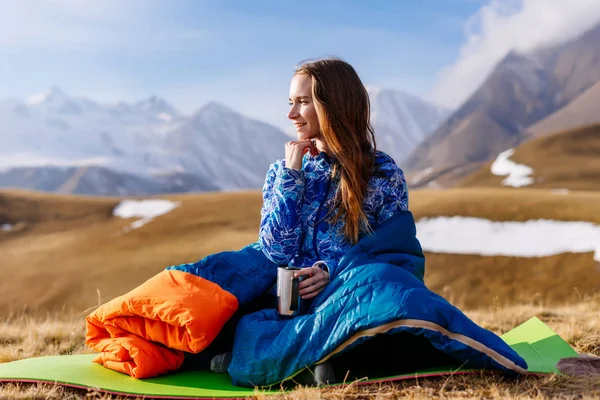 Счастливая длинноволосая девушка сидит в спальном мешке на фоне Кавказских гор, путешествует — стоковое фото