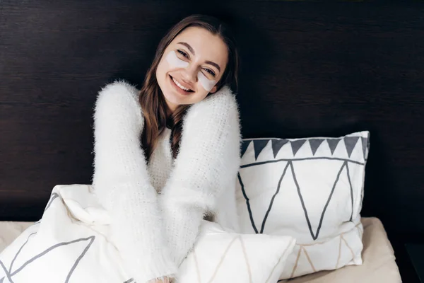 Fröhliches junges Mädchen in weißer Jacke wacht frühmorgens auf, sitzt im Bett, unter den Augen Flecken gegen Schwellungen, lächelt — Stockfoto