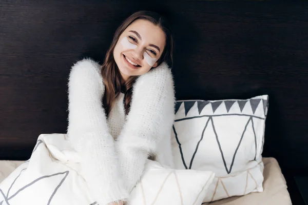 Heureuse jeune fille dans une veste blanche se réveille tôt le matin, s'assoit au lit, sous les yeux patches contre l'enflure, souriant — Photo