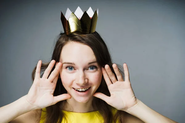 Divertida linda chica en un vestido amarillo sonríe, una corona de oro en su cabeza — Foto de Stock