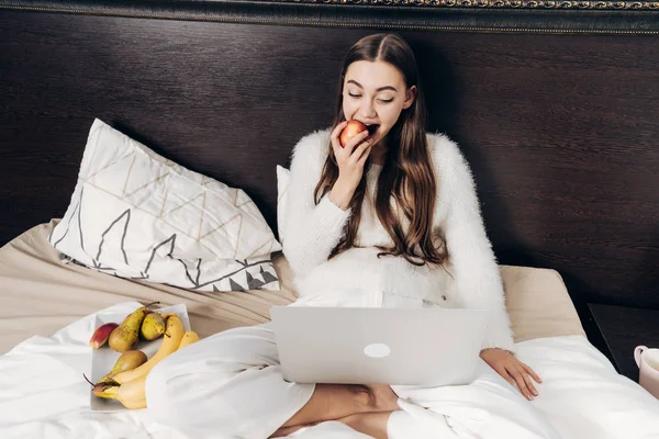 Linda chica de pelo largo en pijama sentado en la cama, comer frutas y ver series de televisión en el ordenador portátil — Foto de Stock