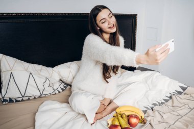 genç kız yatakta bir tabak meyve, yanında oturan bir sabah fotoğraf yapıyor gülümseyen