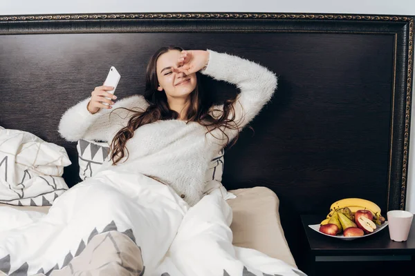 Cute sleepy young girl lies in bed, wakes up early in the morning, stretches, holds smartphone — Stock Photo, Image