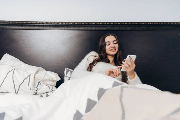 Hermosa chica feliz se encuentra en la cama temprano en la mañana, mira en su teléfono inteligente y sonríe — Foto de Stock