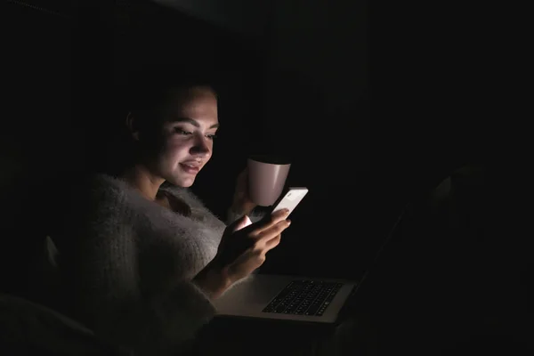 Carina ragazza si siede al buio di notte con un computer portatile, guarda nel suo smartphone e beve il tè — Foto Stock