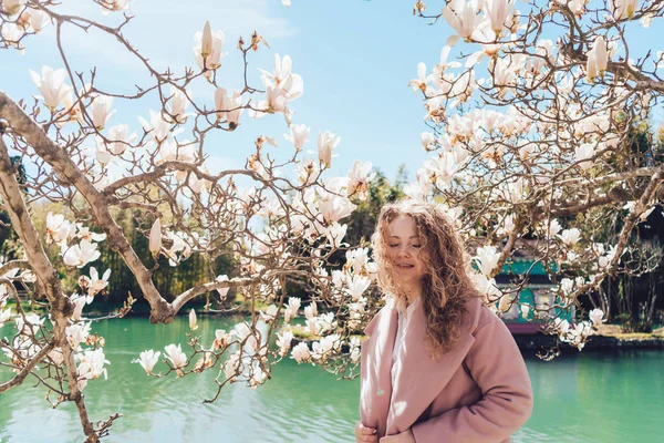 A lovely curly-haired young woman in a pink coat walks the park next to the pond, enjoys the warm spring weather and fragrant magnolia — Stock Photo, Image