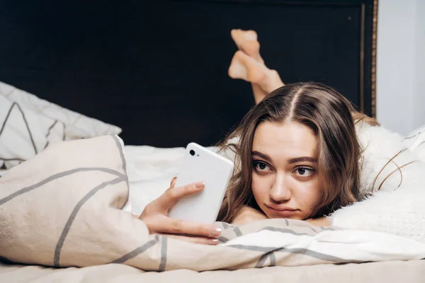 Gadis muda yang lucu berbaring di tempat tidur, beristirahat setelah bekerja keras, melihat ke dalam smartphone-nya — Stok Foto