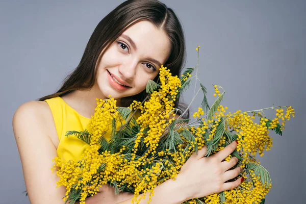 A smiling young girl in a yellow dress is holding a large bouquet of yellow mimosa — Stock Photo, Image