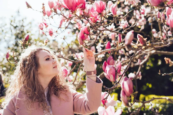 Mujer joven y rizada caminando por el parque de primavera, disfrutando del aroma de la magnolia rosa — Foto de Stock