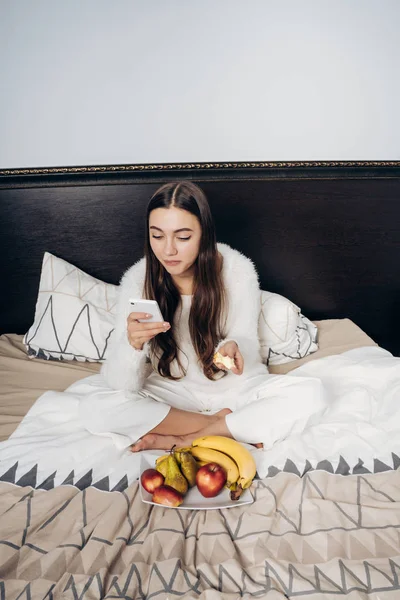Linda chica de pelo largo en pijama blanco comiendo fruta en la cama y mirando en su teléfono inteligente — Foto de Stock
