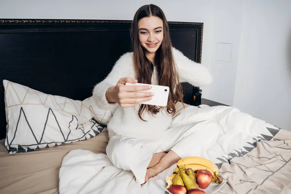 Schöne langhaarige Mädchen sitzt im Bett, frühstückt Obst und macht Selfie — Stockfoto