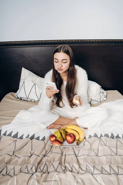 Chica joven de pelo largo en pijama comiendo fruta en la cama y mirando en su teléfono inteligente — Foto de Stock