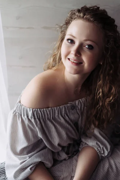 Młoda kobieta kręcone w pięknej sukni siedzi przy oknie, uśmiechając się i patrząc na kamery — Zdjęcie stockowe