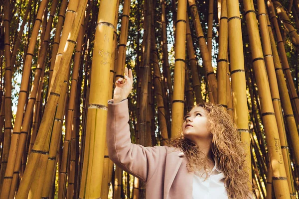 Stylish curly woman in a pink coat walking in the park, looking at bamboo trees — Stock Photo, Image