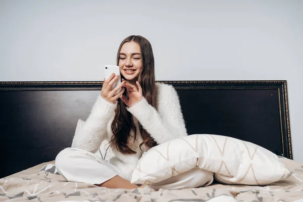 Mooie langharige meisje zit in bed, vroeg in de ochtend wakker, kijkt naar haar telefoon en lacht — Stockfoto