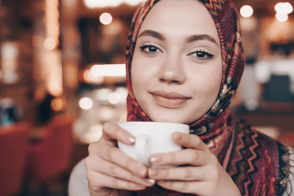 Красивая арабская девушка в восточном шарфе пьет ароматный кофе в кафе и улыбается — стоковое фото